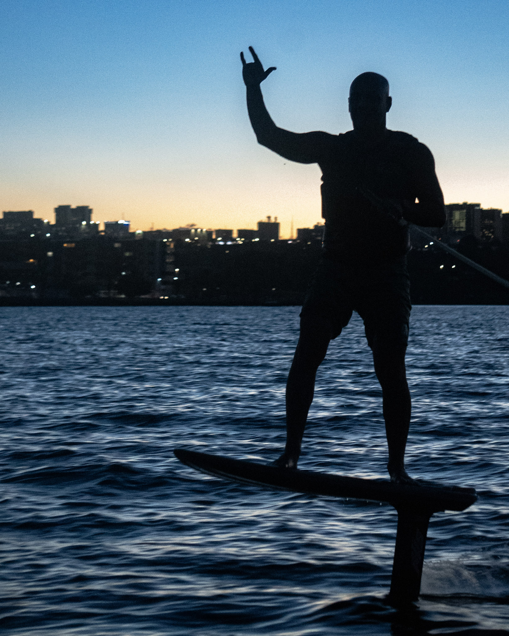 Silhueta de rider sobre o foil ao anoitecer, com as luzes da cidade ao fundo