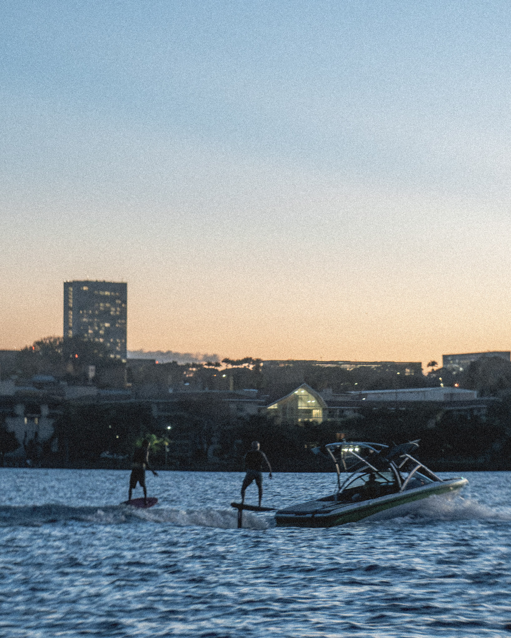 Dois riders de foil e a lancha no fim de tarde do Lago Paranoá, com a cidade ao fundo