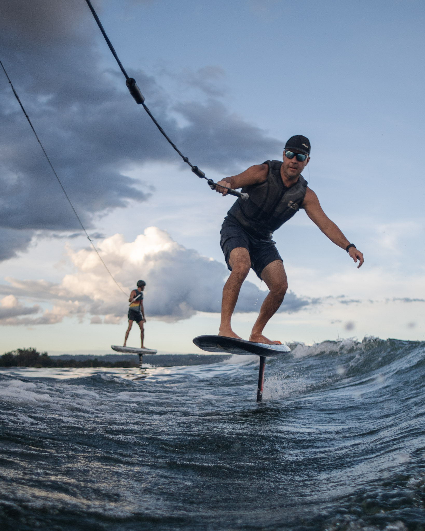Dois riders de wakefoil voando sobre a água do Lago Paranoá, um deles puxado pela corda