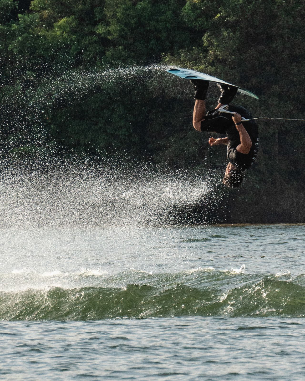Wakeboarder invertido em pleno ar sobre a onda, com a mata do lago ao fundo